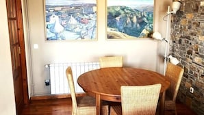 A dining area with a round wooden table and wicker chairs, two paintings on the wall, and a stone wall on one side. - Townhouse 4 rooms Pool, Fireplace, Wi-Fi & Pet-Friendly (Llívia)