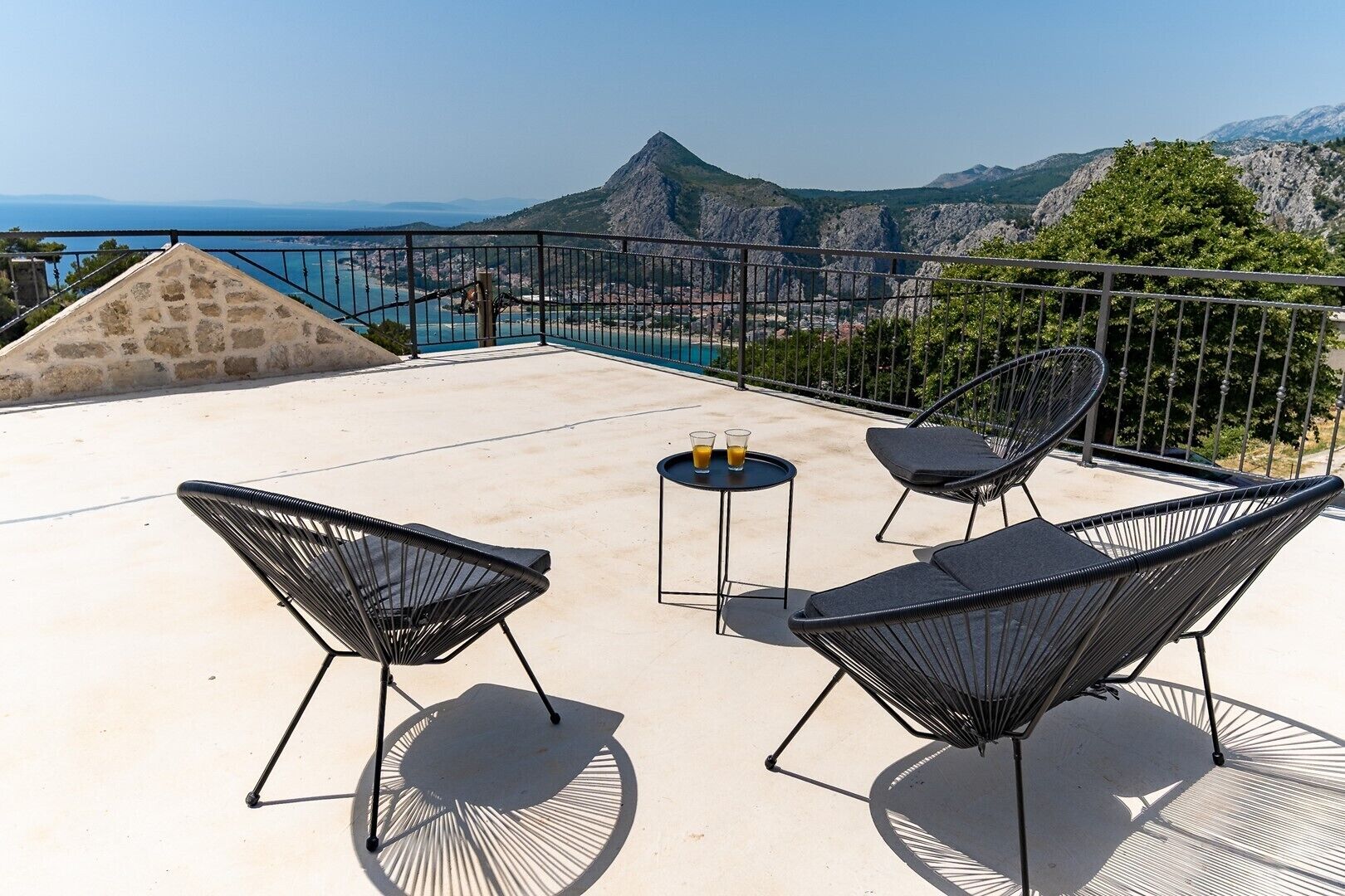 Een dakterras met vier zwarte frame stoelen, een kleine ronde tafel met twee glaasjes sinaasappelsap, en uitzicht op een bergachtige kustlijn.