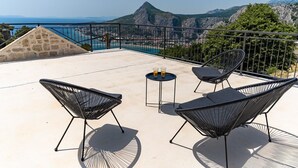 A rooftop terrace with four black wireframe chairs, a small round table with two glasses of orange juice, and a view of a mountainous coastline. - Seaview Villa KING with pool 3km from sea (Omis)
