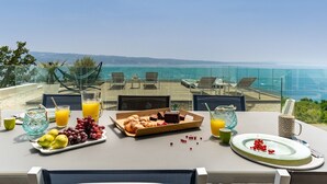 Una mesa dispuesta con alimentos para el desayuno, como frutas, pasteles y bebidas, con vistas a un hermoso panorama marino.