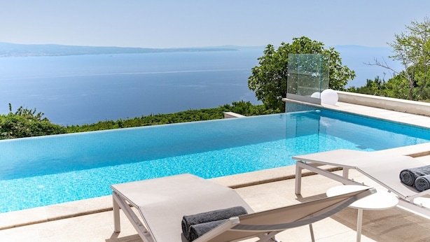 Una piscina infinita azul cristalina con vistas al mar, sillas de descanso y un área con paredes de vidrio.