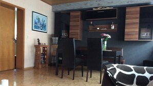 A living room with a sofa, chairs, a coffee table, and a dining area with a table and chairs. There is a painting on the wall and a vase with flowers on the dining table.