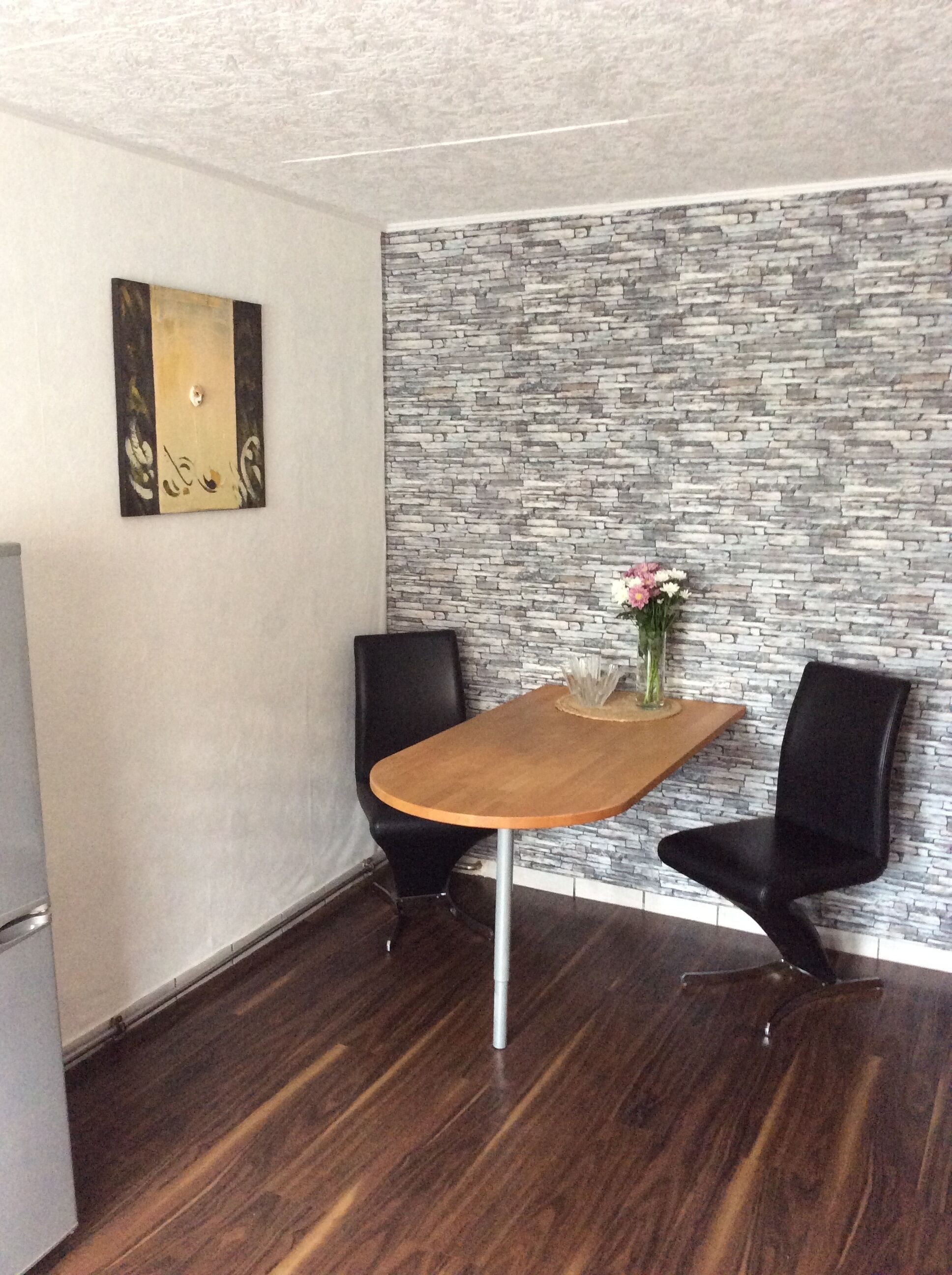 A dining area with a wooden table, two black chairs, a vase with flowers, and a stone-patterned wall.