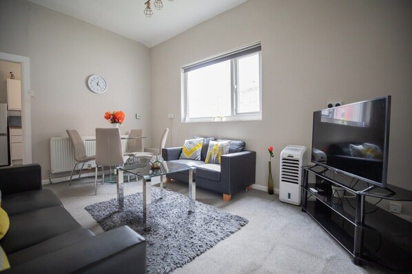 A modern living room with a grey sofa, a glass coffee table, a grey rug, a TV on a stand, and a window with blinds.