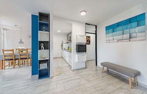 Een moderne keuken met witte kastjes, een ingebouwde oven en een televisie. Er is een eethoek met houten stoelen en een tafel, en een bankje in de hoek.