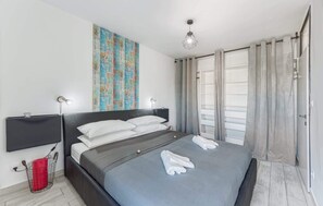 Una habitación de hotel moderna con una cama grande, una mesita de noche, un cesto de basura rojo y un televisor montado en la pared.