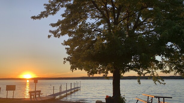 A sunset view over a lake with a wooden dock, picnic tables, and a stone wall. - Lakeview Cabin- minutes from beach! (Crofton)
