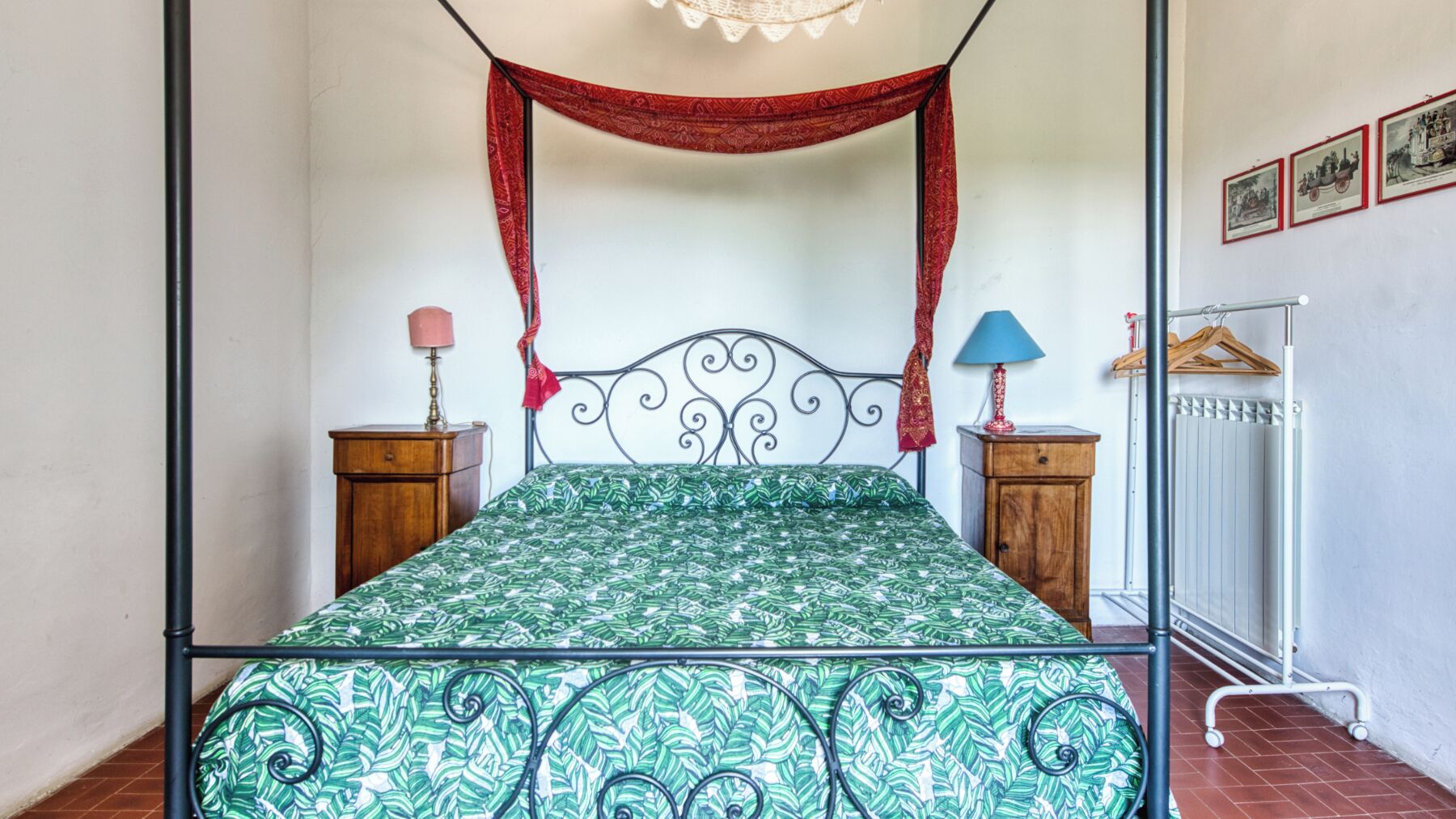 A four-poster bed with a green patterned bedspread, two bedside tables with lamps, a radiator, and framed pictures on the wall.