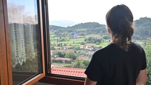 Une personne se tient près d’une fenêtre, d’où l’on peut admirer un paysage rural, avec des champs et des maisons.