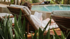 Zona de piscina con sillones de descanso, una persona leyendo un libro y abundante vegetación.