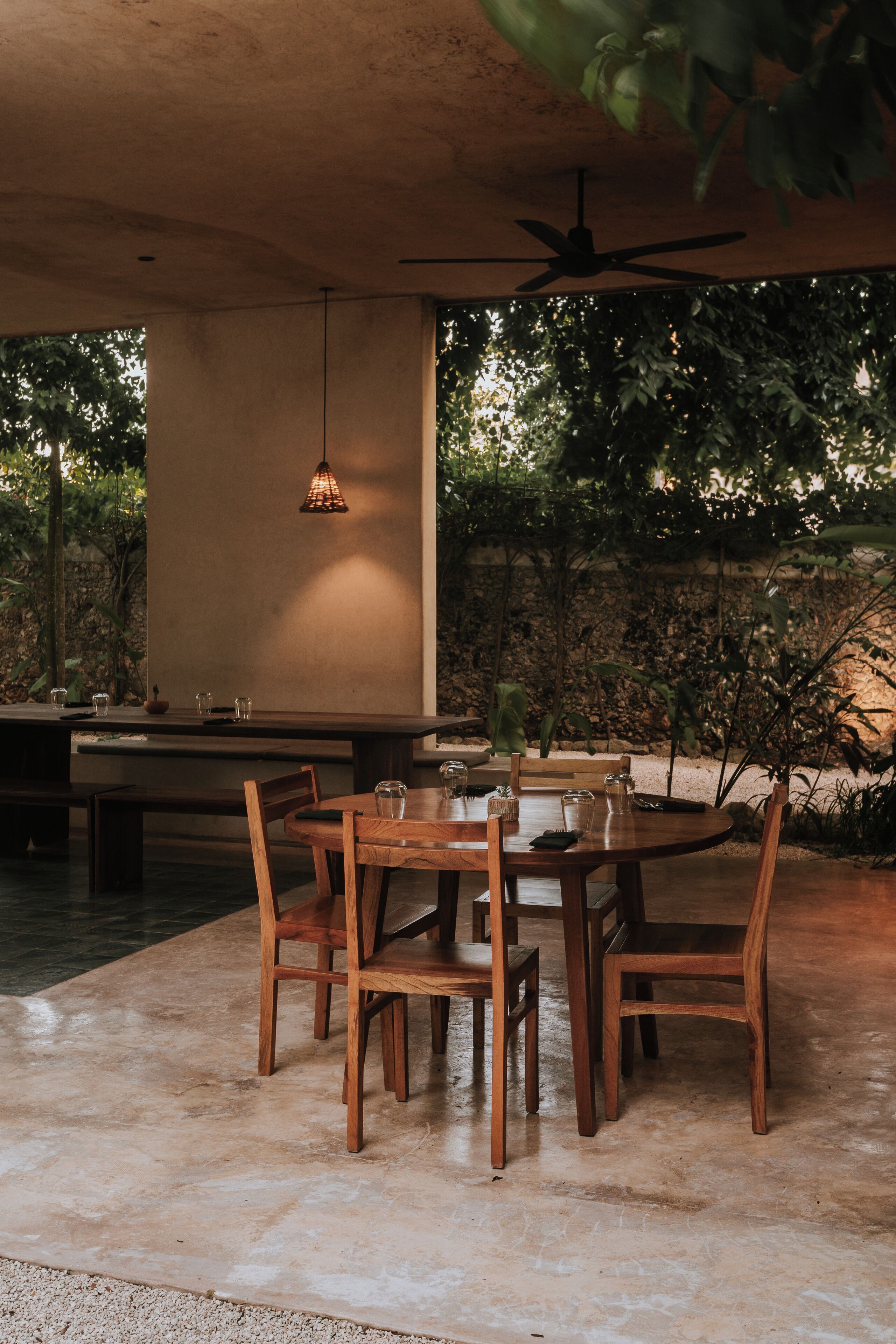 Un espacio de comedor al aire libre con una mesa redonda y sillas de madera, rodeado de follaje y un ventilador de techo.