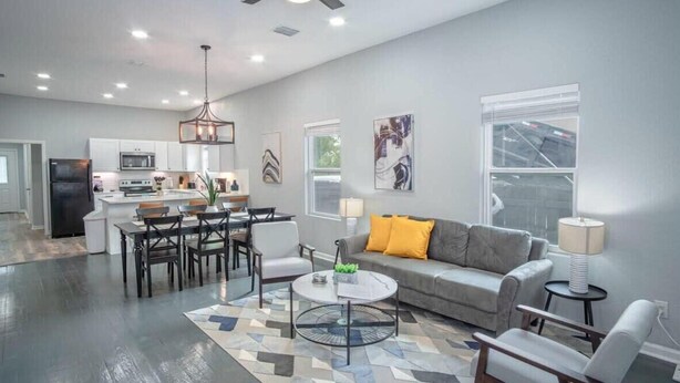 A modern kitchen with white cabinets, a dining area with a round table and chairs, a gray sofa with yellow cushions, and a ceiling fan.