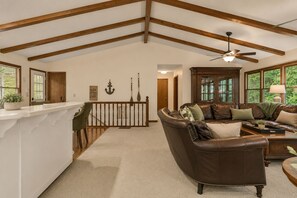 A spacious living area with a high ceiling, a wooden ceiling beam structure, a brown leather sofa, a dining area with a white counter, and a view of greenery through large windows.