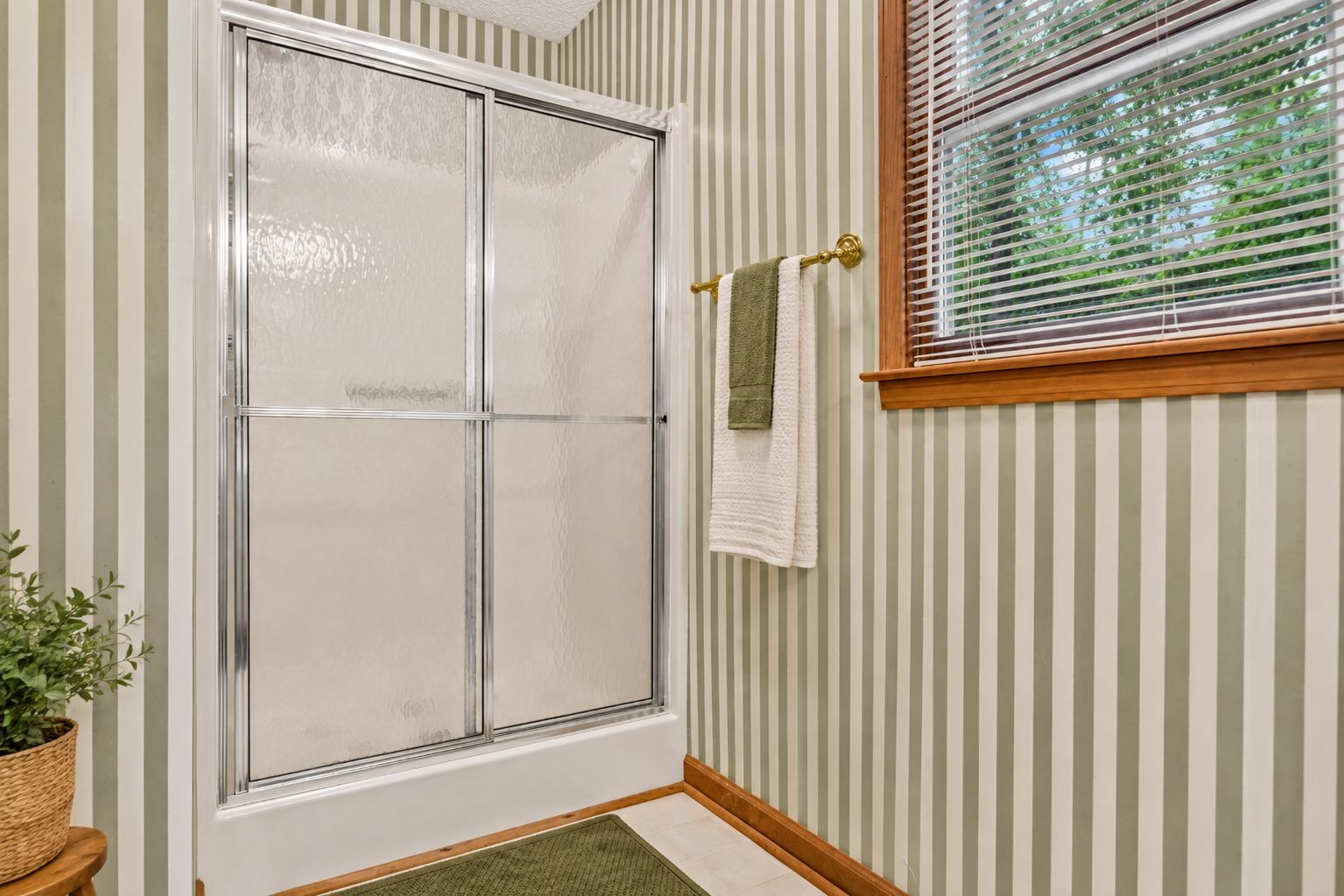 A bathroom with a glass shower door, striped wallpaper, a window with blinds, and a towel rack with two towels.