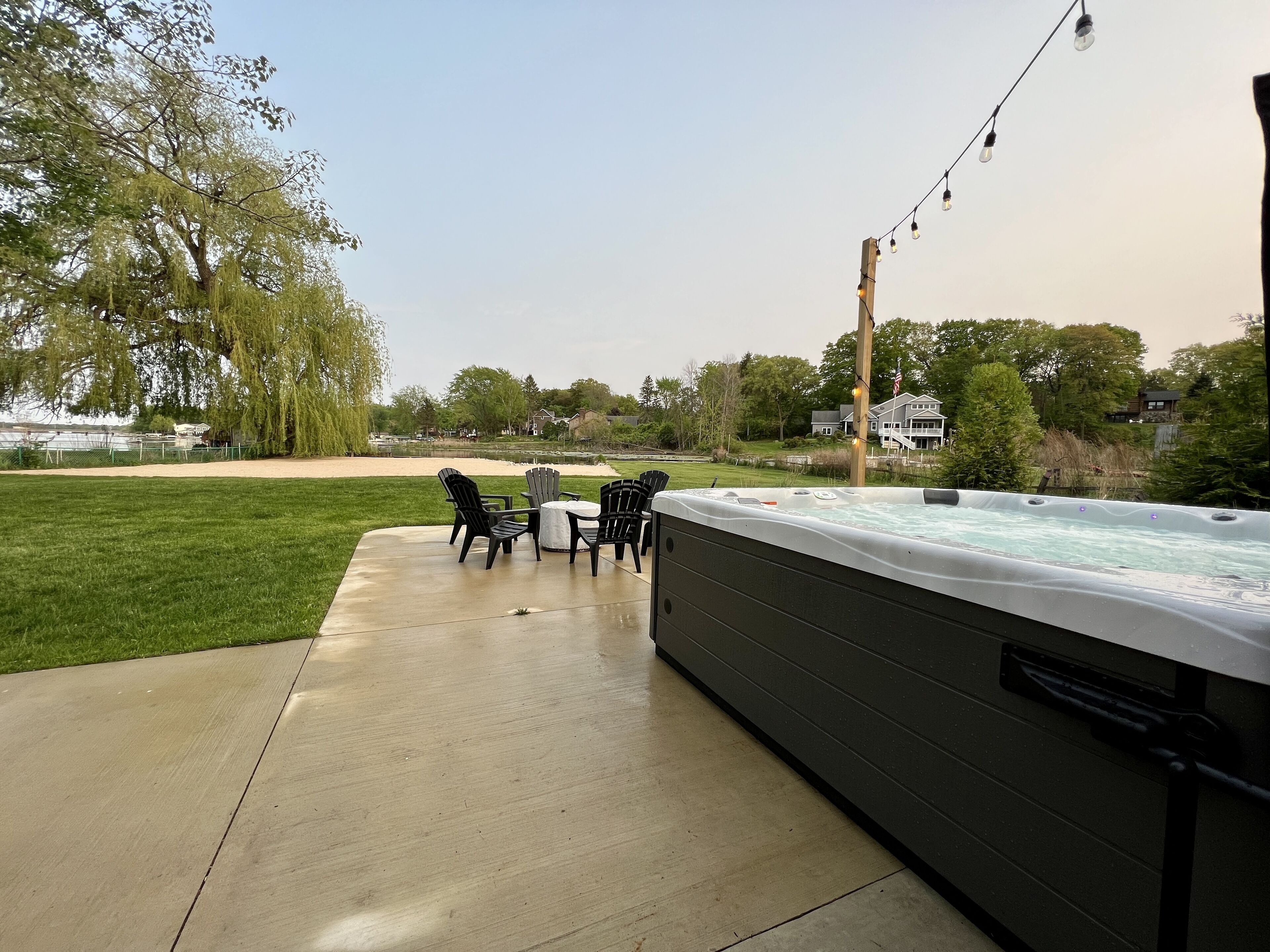 A hot tub with four Adirondack chairs and a table, surrounded by a grassy yard and a wooden deck.