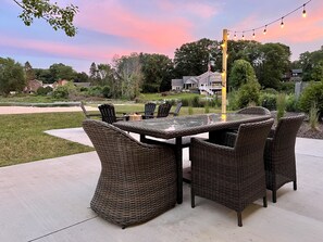 An outdoor dining area with wicker furniture and a glass-top table, surrounded by a concrete patio and a view of a residential neighborhood. - Lakefront Home - Hot Tub & Private Beach (Spring Lake)