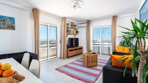 A modern living room with a dining area, a black sofa with orange cushions, a wooden TV stand, and a large window offering a city view. - Faro Center Stunning View (Faro)
