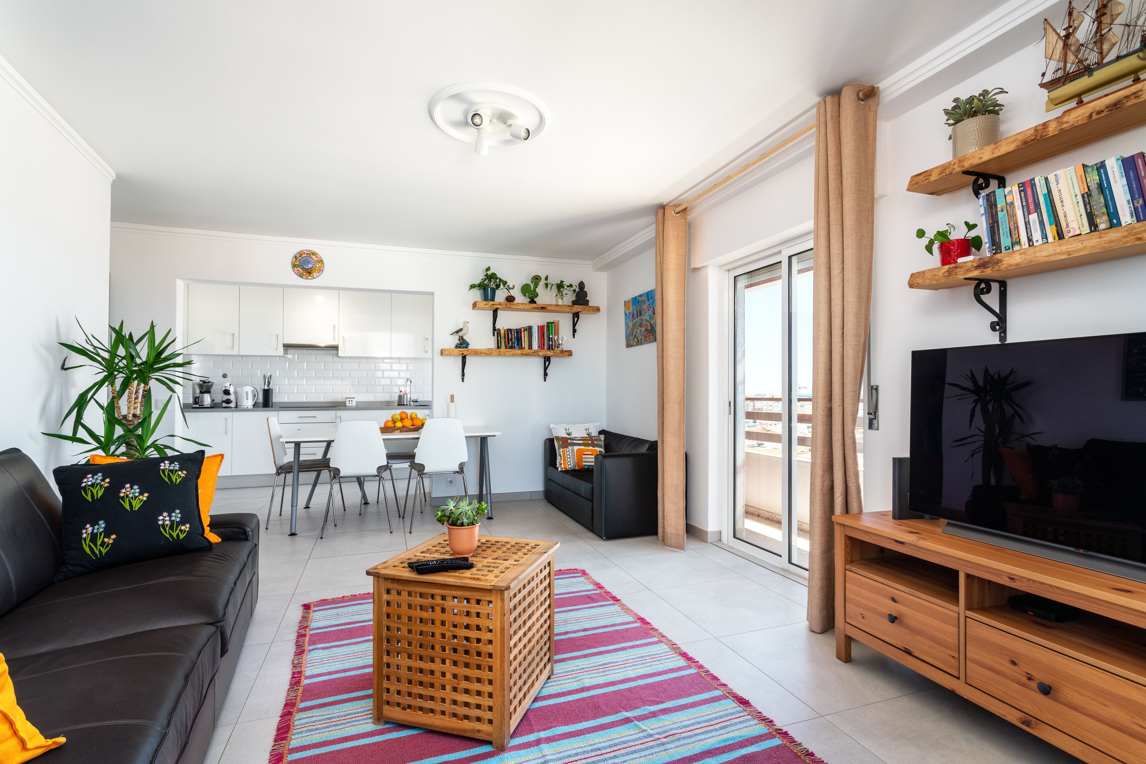 A modern living room with a black sofa, a wooden coffee table, and a TV stand. There is a kitchen in the background with white cabinets and a dining area with a table and chairs.