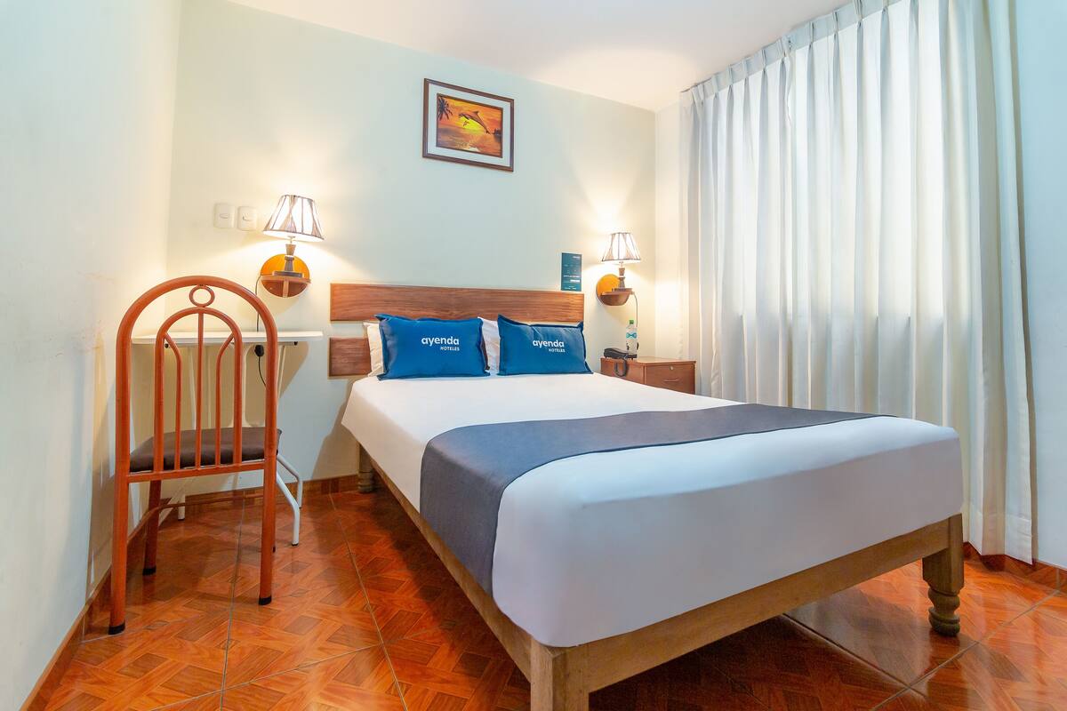 Ein Hotelzimmer mit einem hölzernen Bett, zwei blauen Kissen, einem Holzstuhl, einem Nachttisch mit Lampe und einem fenster mit Vorhängen.