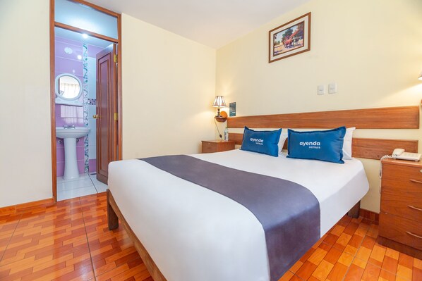 A hotel room with a large bed, wooden headboard, two pillows with the hotel name, a nightstand with a lamp, a wooden dresser, and a bathroom visible through an open door. - Hotel Musuk Wasi (Arequipa)