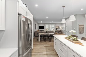 A modern kitchen with a stainless steel refrigerator, a dining area with a wooden table and chairs, and a living room with a sofa and a TV. - New Saugatuck Home: Lake Views & Walk to Town (Douglas)