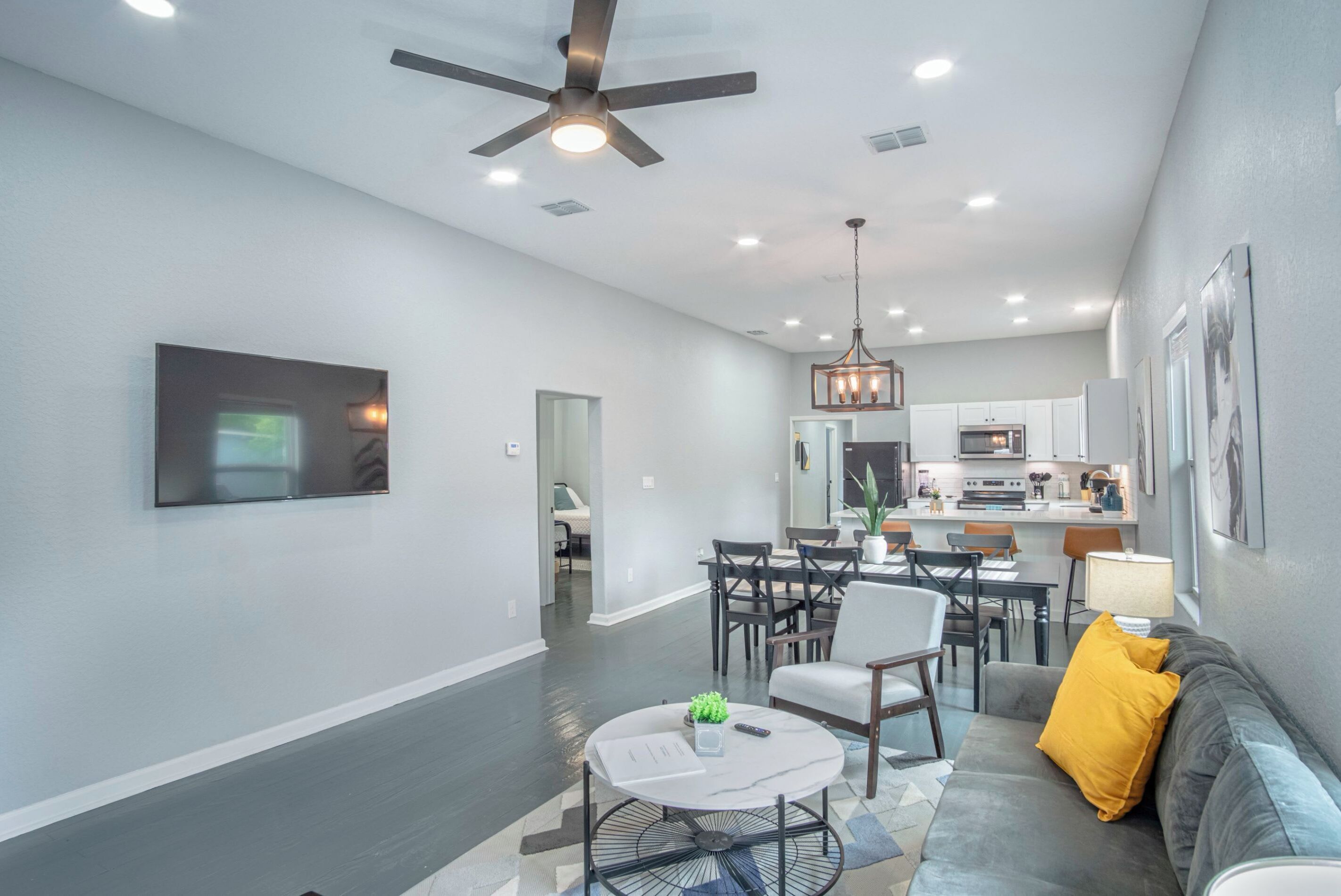 A spacious living area with a ceiling fan, a dining table with chairs, a sofa with cushions, and a kitchen in the background.