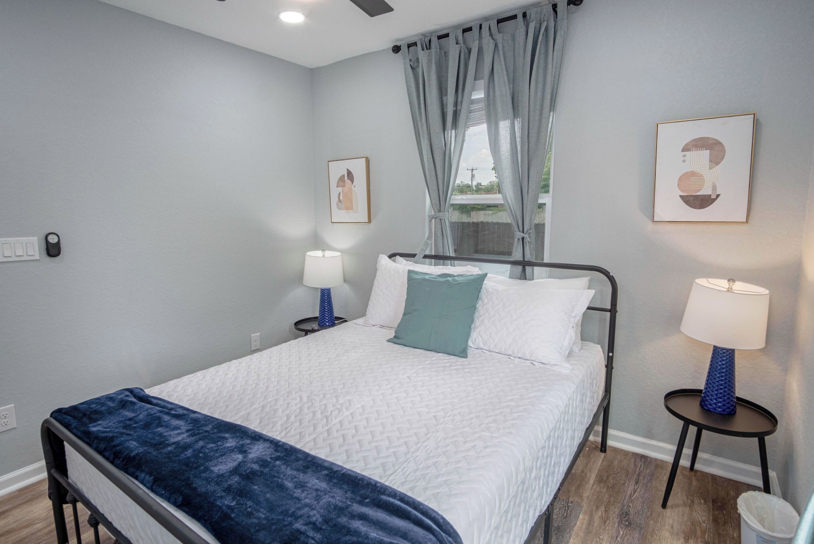 Un dormitorio ordenado con una cama, dos mesitas de noche con lámparas, una ventana con cortinas y dos cuadros enmarcados en la pared.