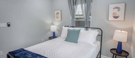 Un dormitorio ordenado con una cama, dos mesitas de noche con lámparas, una ventana con cortinas y dos cuadros enmarcados en la pared.