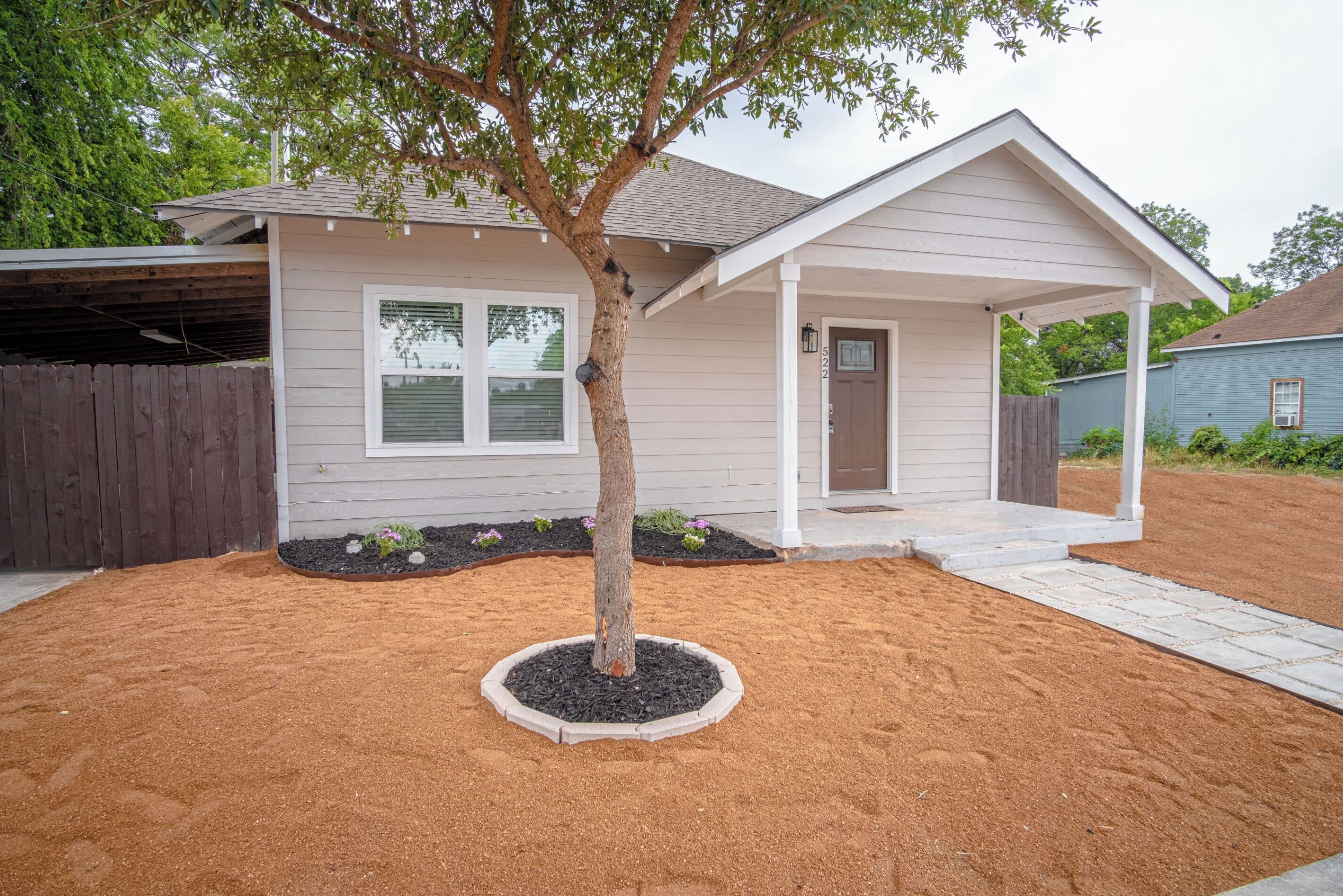 A residential house with a covered porch, a tree in the front yard, and a sandy driveway.