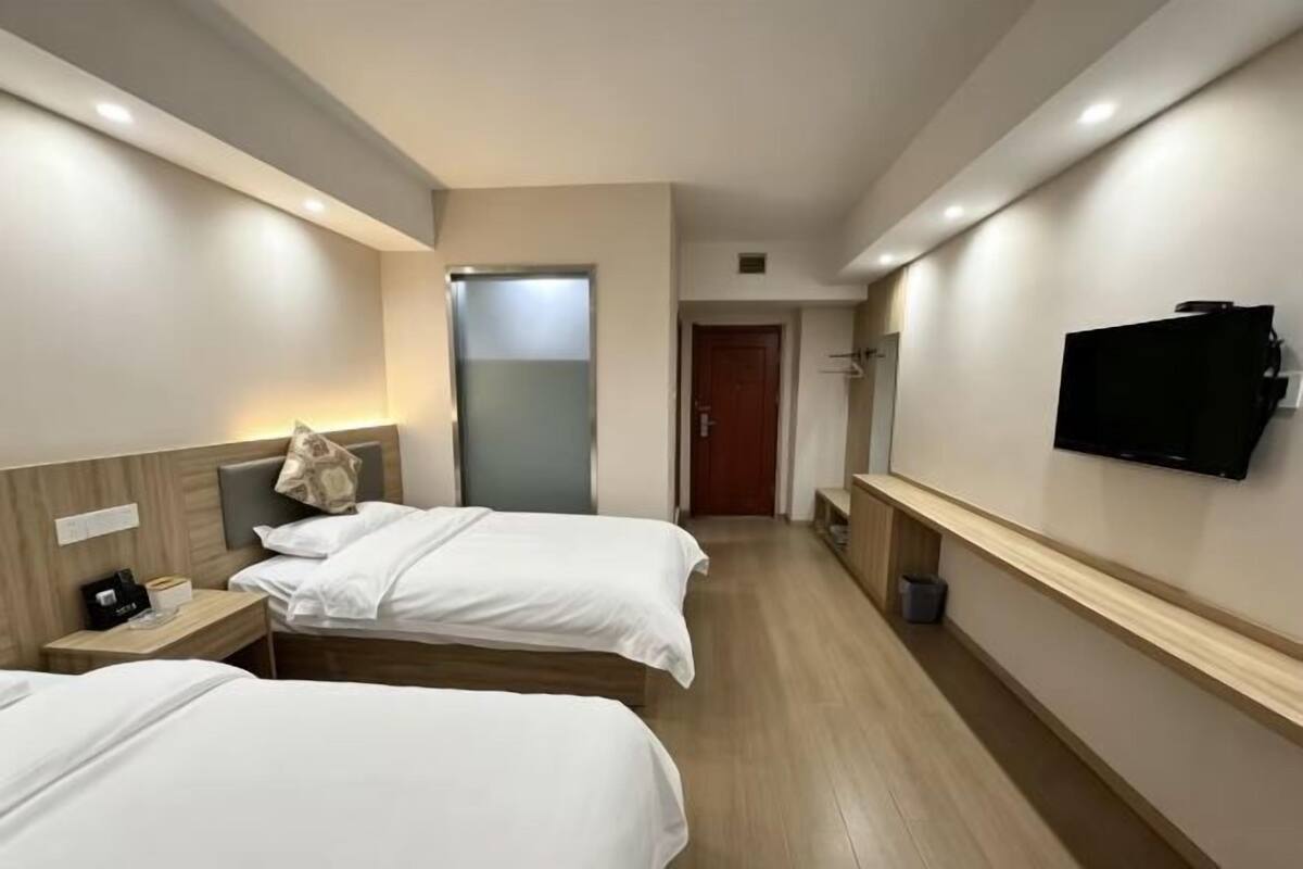 Ein Hotelzimmer mit zwei Betten, einem an der Wand montierten Fernseher, einem hölzernen Kopfteil und einem kleinen Nachttisch.