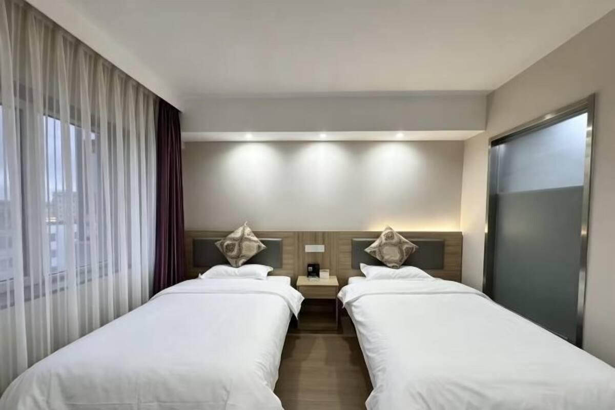 Ein Hotelzimmer mit zwei Betten, einem Nachttisch, einer Lampe und einem großen Fenster mit Vorhängen.