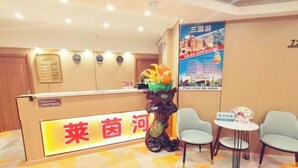 A hotel reception area with a reception desk, a decorative plant, two chairs, a small table, and a poster on the wall.