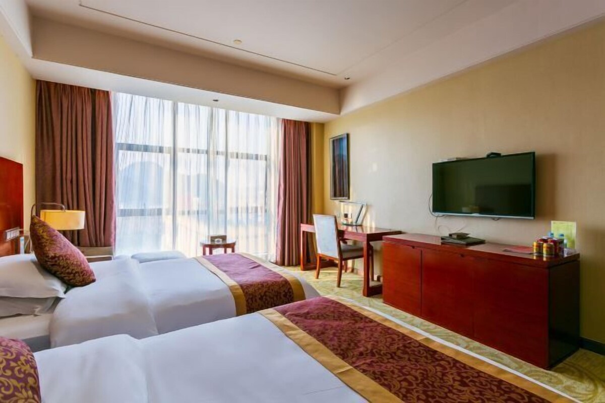 A hotel room with two beds, a desk, a television, and a large window with curtains.