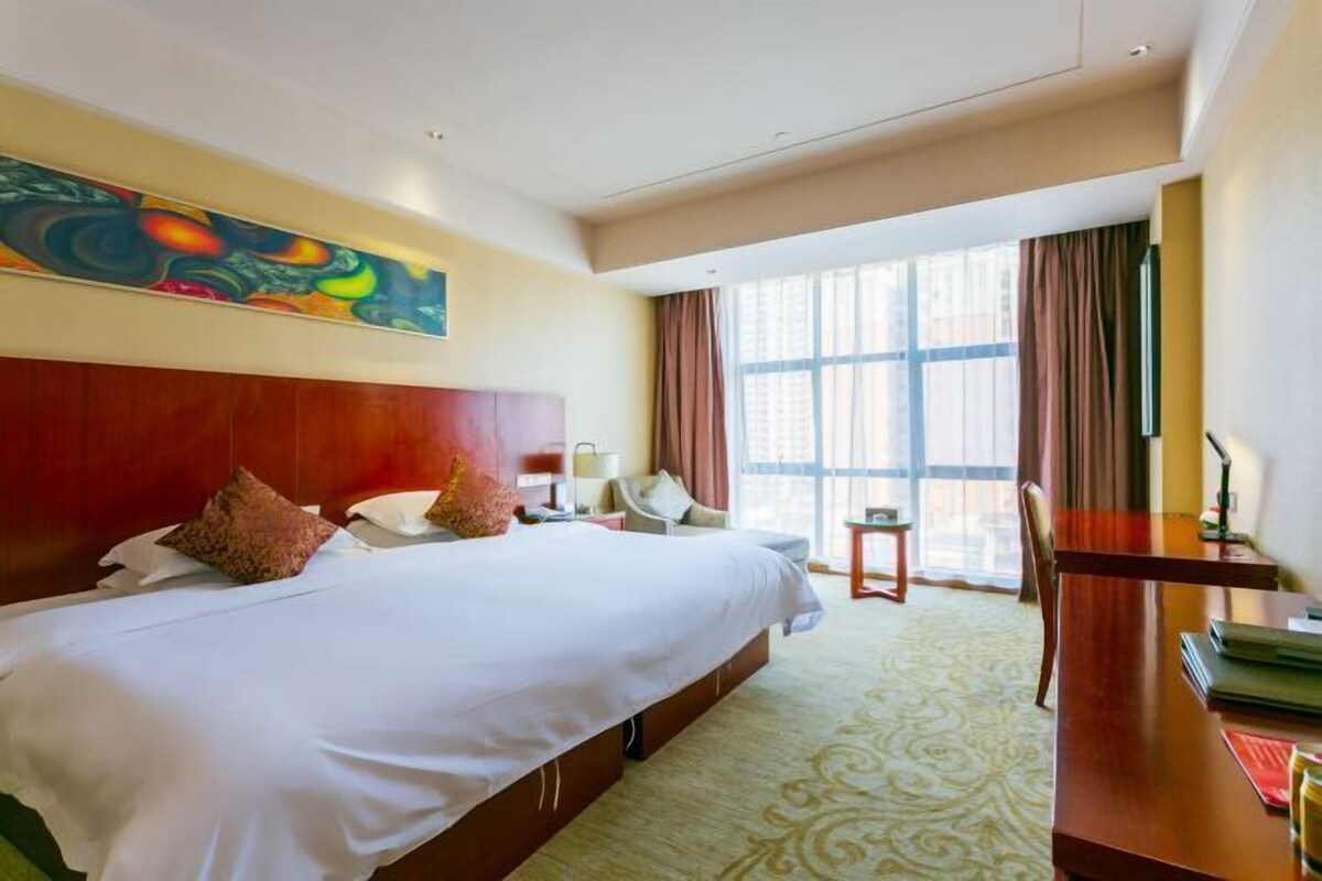 A hotel room with a large bed, a desk, a chair, a window with curtains, and a colorful painting on the wall.