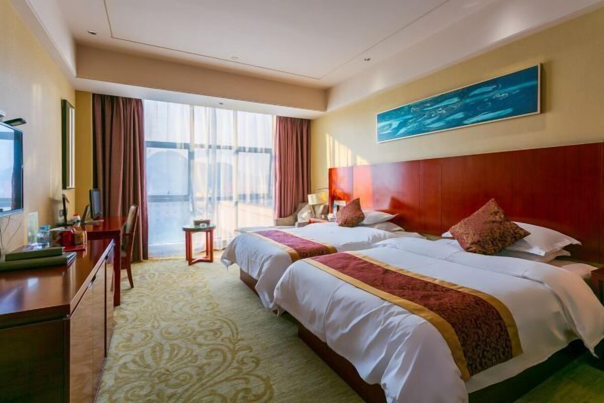 A hotel room with two beds, a desk, a chair, a TV, and a large window with curtains.