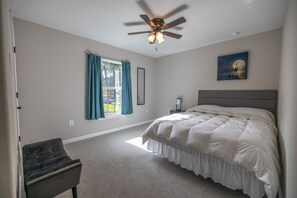 A bedroom with a bed, a ceiling fan, a window with curtains, a nightstand, and a painting on the wall. - Cheerful 3 bedroom home with pool + grill (Lakeland)