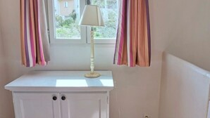 A room with a white cabinet, a lamp on top, and a window with curtains. - 45 m2 house, facing the pine forest, terrace, garden and tranquillity (Mallemort)