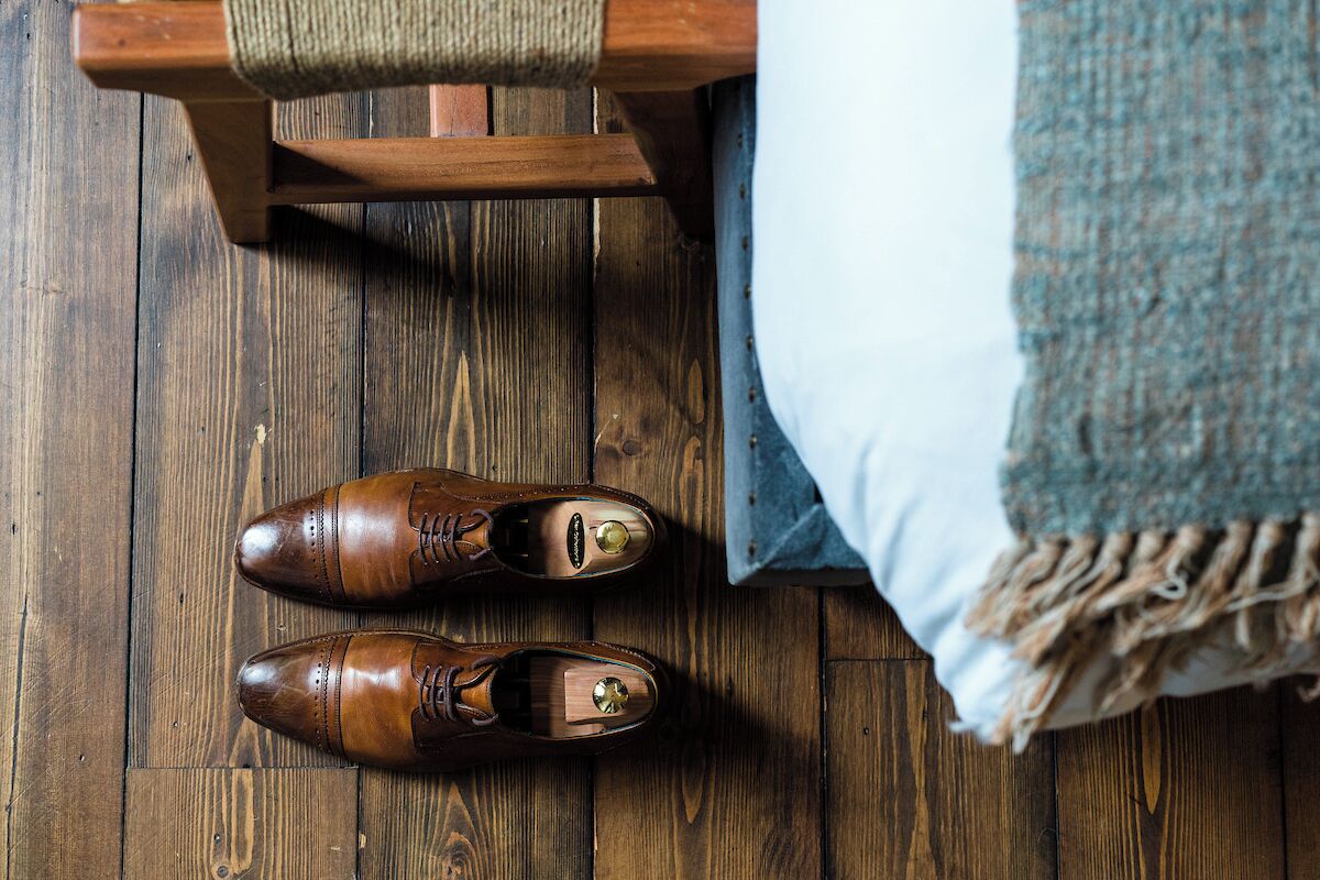 A pair of brown leather shoes on a wooden floor next to a bed with a blue blanket.
