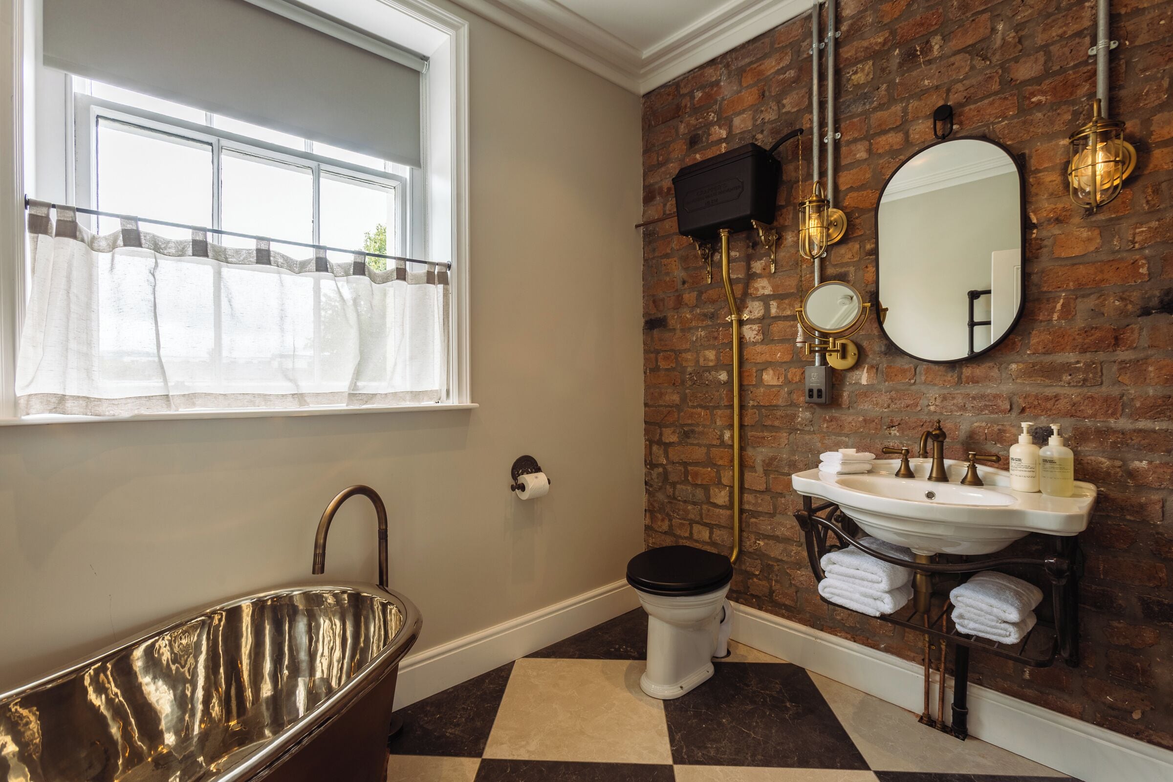 A bathroom with a freestanding bathtub, a toilet, a sink with a mirror, and a brick wall.