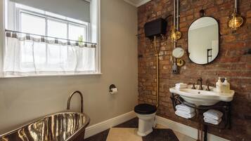 A bathroom with a freestanding bathtub, a toilet, a sink with a mirror, and a brick wall.