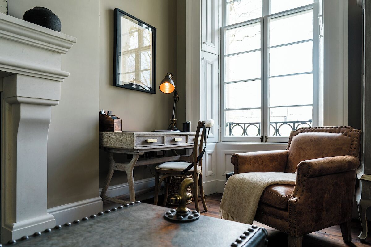 A room with a fireplace, a desk with a lamp, a leather armchair, and a large window with a view.