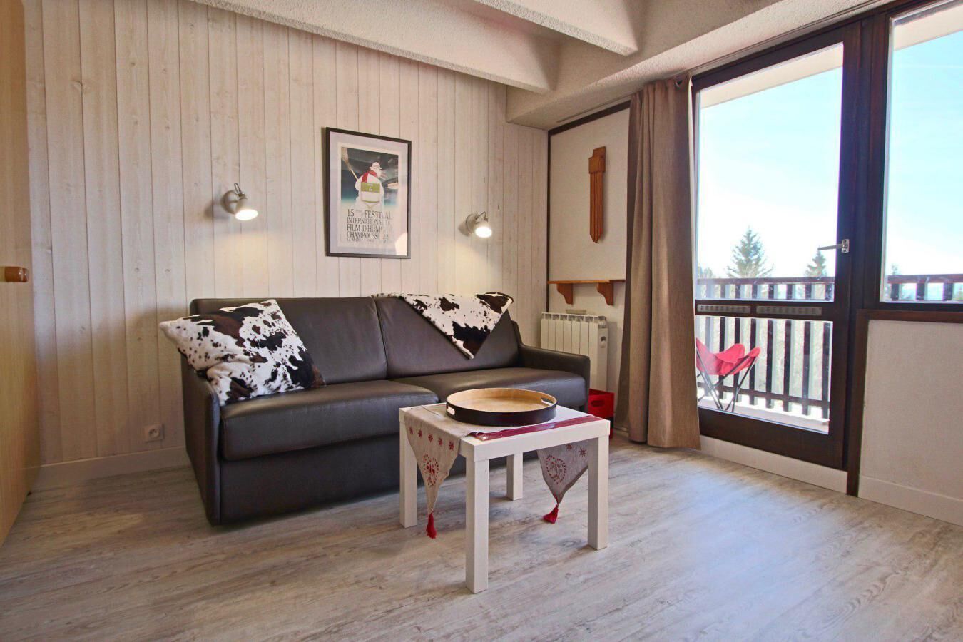 Un salon chaleureux avec un canapé en cuir foncé, une petite table en bois sur laquelle repose un plateau, et une grande fenêtre donnant sur un balcon.