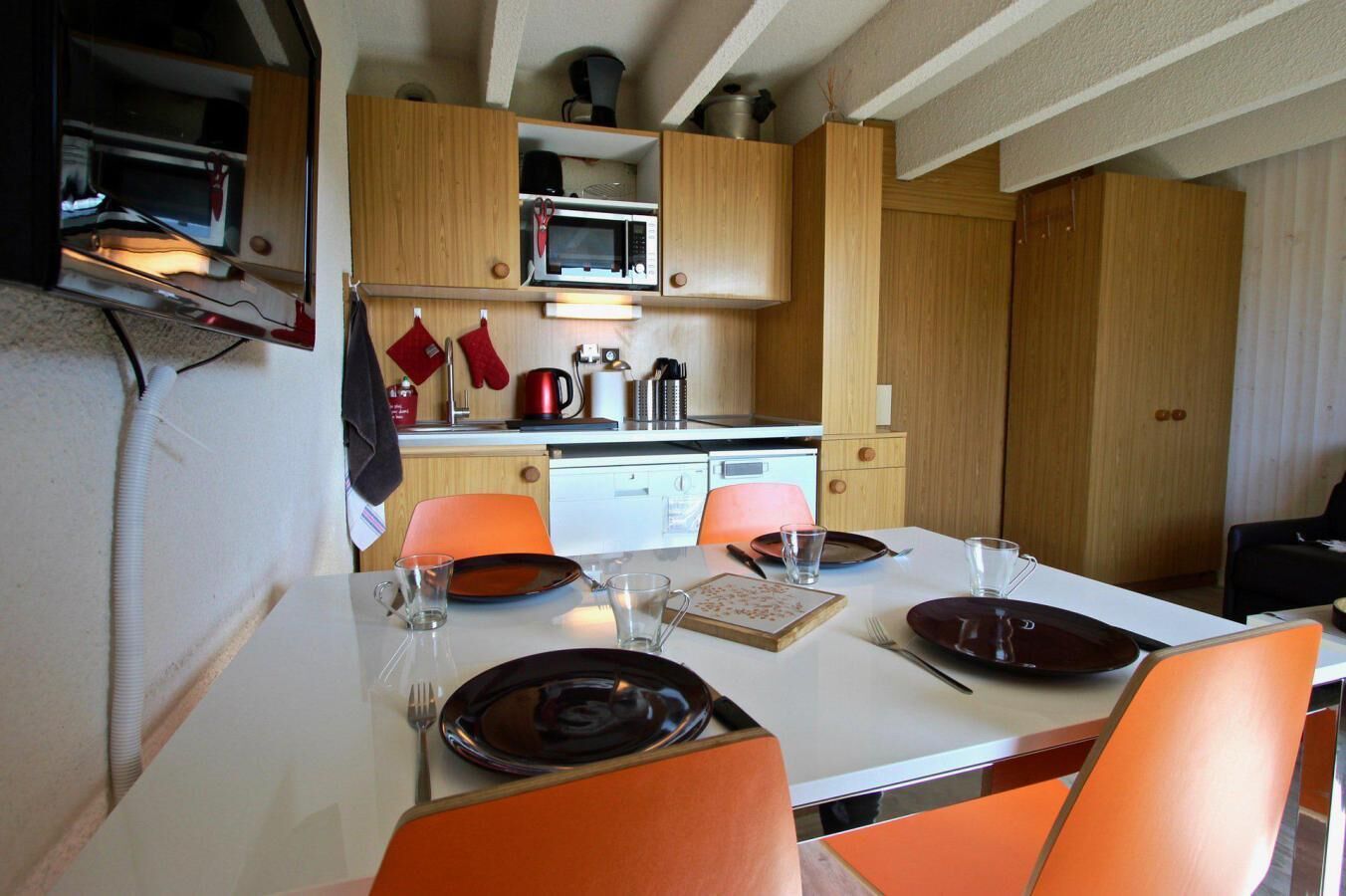 A compact kitchen with wooden cabinets, a microwave, and a coffee maker. There is a dining table with orange chairs and a white tablecloth.