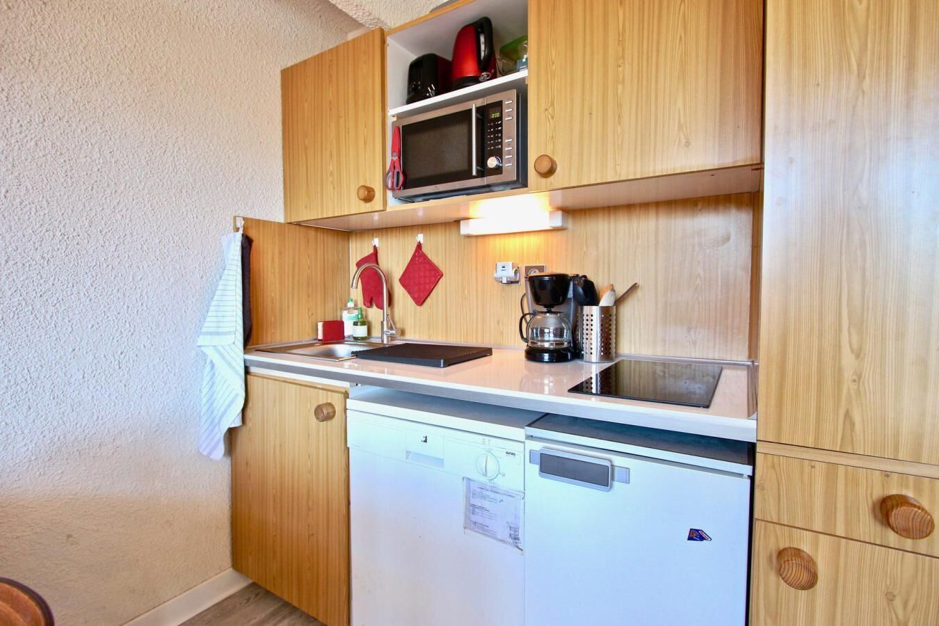 Cocina compacta con armarios de madera, microondas, cafetera y lavavajillas.