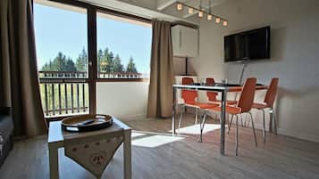 Una habitación con balcón, una mesa de comedor con sillas, un televisor de pantalla plana y una mesita con bandeja y control remoto.