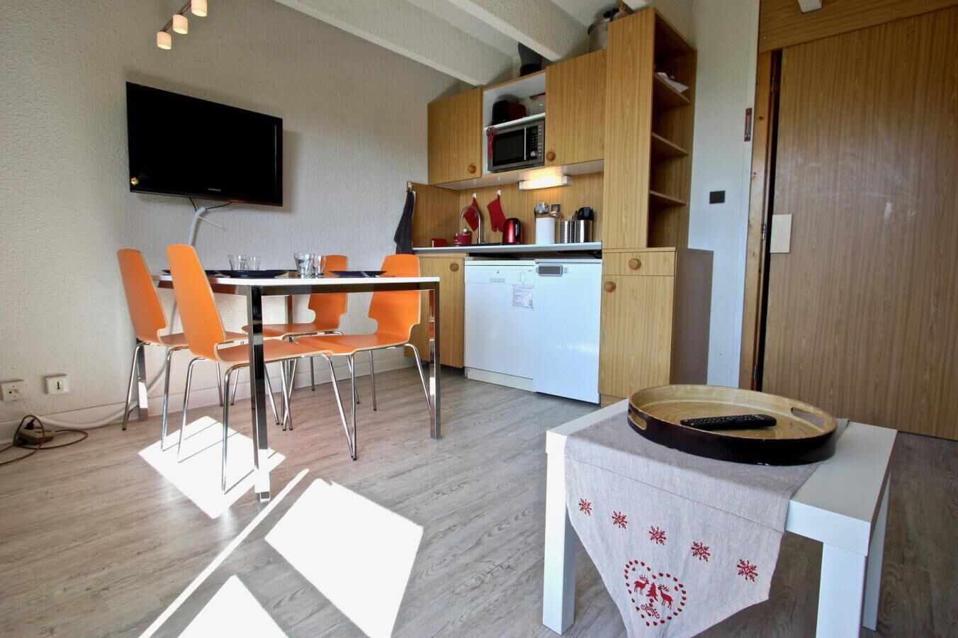 Eine kompakte Küche mit Mikrowelle, Backofen und weißem Geschirrspüler. Ein Essbereich mit orangefarbenen Stühlen und einem Tisch. Ein Flachbildfernseher ist an der Wand montiert.