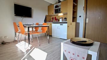 Eine kompakte Küche mit Mikrowelle, Backofen und weißem Geschirrspüler. Ein Essbereich mit orangefarbenen Stühlen und einem Tisch. Ein Flachbildfernseher ist an der Wand montiert.