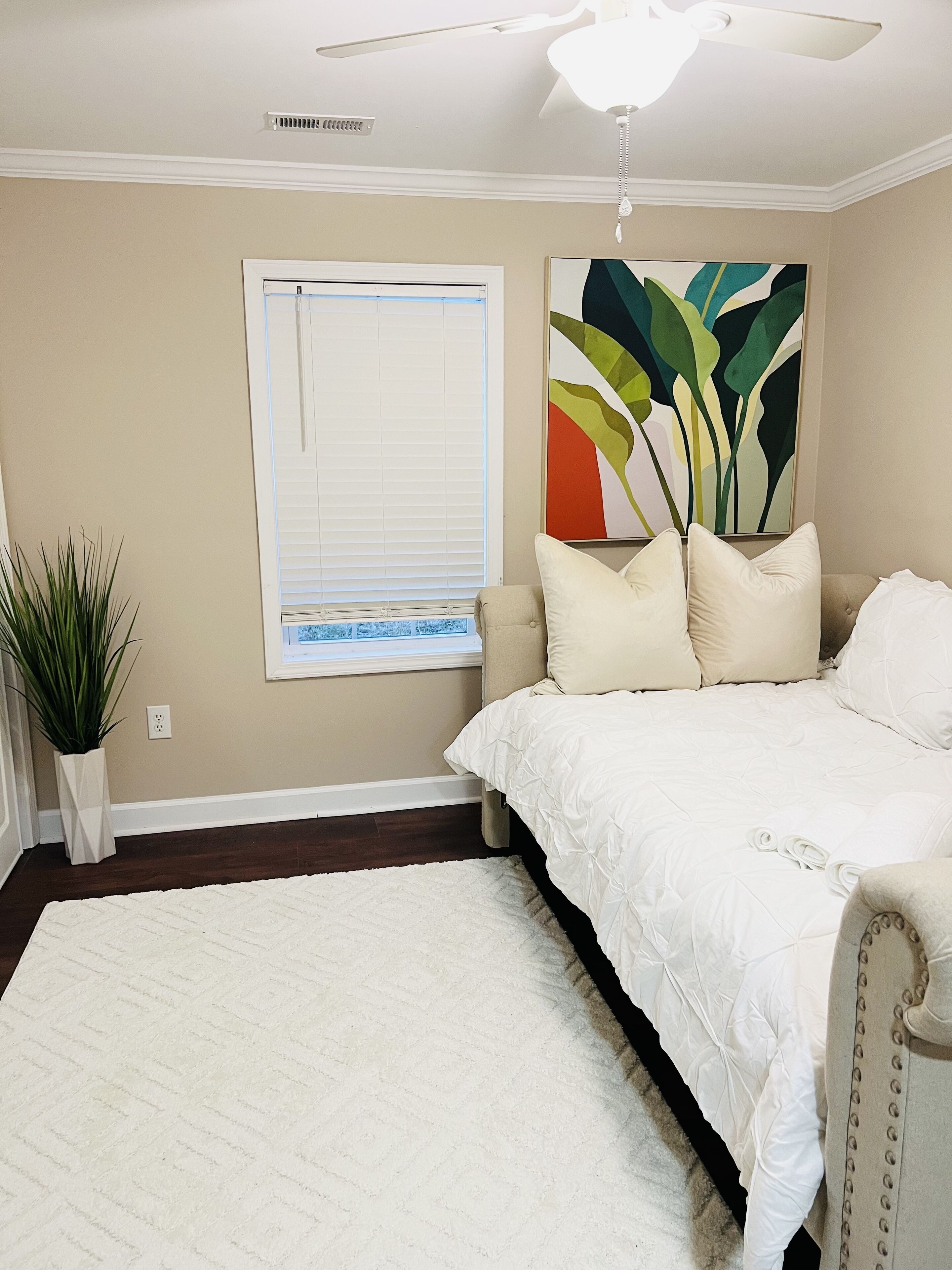 A bedroom with a bed, a ceiling fan, a window with blinds, a painting, and a potted plant.