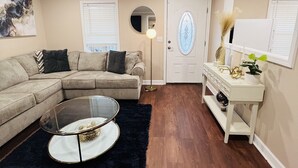 Una sala de estar con un sofá modular, una mesa de centro de vidrio, un tapete oscuro, una consola blanca con objetos decorativos y una puerta con un vitral.