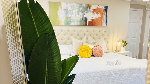 Un dormitorio con cabecera capitonada, cama con ropa de cama blanca, dos almohadas decorativas, una alfombra colorida y una planta en maceta.